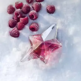 A vibrant pink star-shaped Mugler Angel Nova bottle lying in white snow, surrounded by scattered fresh red raspberries that highlight its fruity notes.