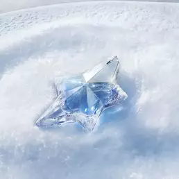 A close-up shot of the iconic light blue, star-shaped Mugler Angel perfume bottle resting elegantly on a bed of pristine, sparkling white snow under soft blue light.