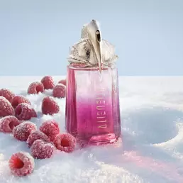 A bright pink Mugler Alien pulp eau de parfum perfume bottle standing upright in the snow, with several fresh red raspberries scattered around its base.