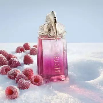 A bright pink Mugler Alien pulp eau de parfum perfume bottle standing upright in the snow, with several fresh red raspberries scattered around its base.