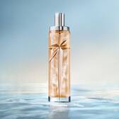 Elegant tall cylindrical perfume bottle with silver cap, filled with warm golden-amber juice, standing on a reflective surface mimicking calm water against a soft light blue background.