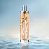 Elegant tall cylindrical perfume bottle with silver cap, filled with warm golden-amber juice, standing on a reflective surface mimicking calm water against a soft light blue background.