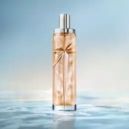 Elegant tall cylindrical perfume bottle with silver cap, filled with warm golden-amber juice, standing on a reflective surface mimicking calm water against a soft light blue background.