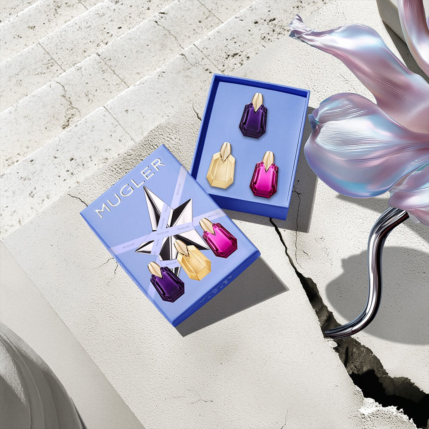 The Mugler Mother's Day 2026 Discovery Set opened to reveal three Alien miniatures (EDP, Goddess, Pulp) in a blue floral box, set on sunlit cracked stone.