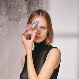 Portrait of a woman with short blonde hair holding the nude-toned Angel Blush star bottle in front of her eye, embodying pure glamour and an intimate glow.