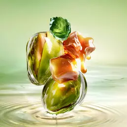 New Starlicious Pistachio Praline still life image featuring the slender bottle surrounded by its key ingredients: a patchouli leaf, whole pistachios, and dripping caramel cubes, all set against a luminous green rippling water background.