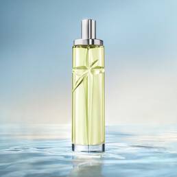 An elegant, tall, cylindrical perfume bottle with a silver cap stands vertically against a soft, light blue background. The bottle is filled with a pale green juice, and it sits on a reflective surface that mimics the ripples of clear, calm water, with the horizon line softly blurred in the background.