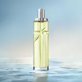 An elegant, tall, cylindrical perfume bottle with a silver cap stands vertically against a soft, light blue background. The bottle is filled with a pale green juice, and it sits on a reflective surface that mimics the ripples of clear, calm water, with the horizon line softly blurred in the background.