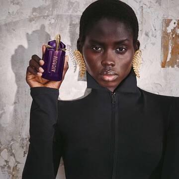 Person holding Mugler Alien purple perfume bottle against textured wall