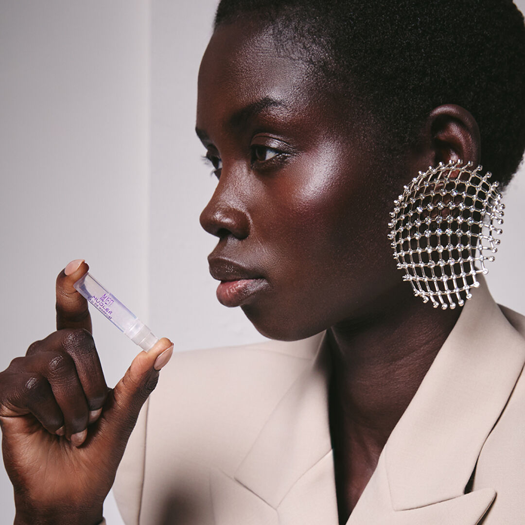 Close-up of a model with a silver ear cuff holding three Mugler fragrance travel sprays, including Angel and Alien, against a textured industrial wall.