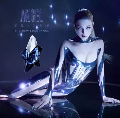 Hunter Schafer in a metallic body suit posing next to a bottle of Mugler Angel Elixir