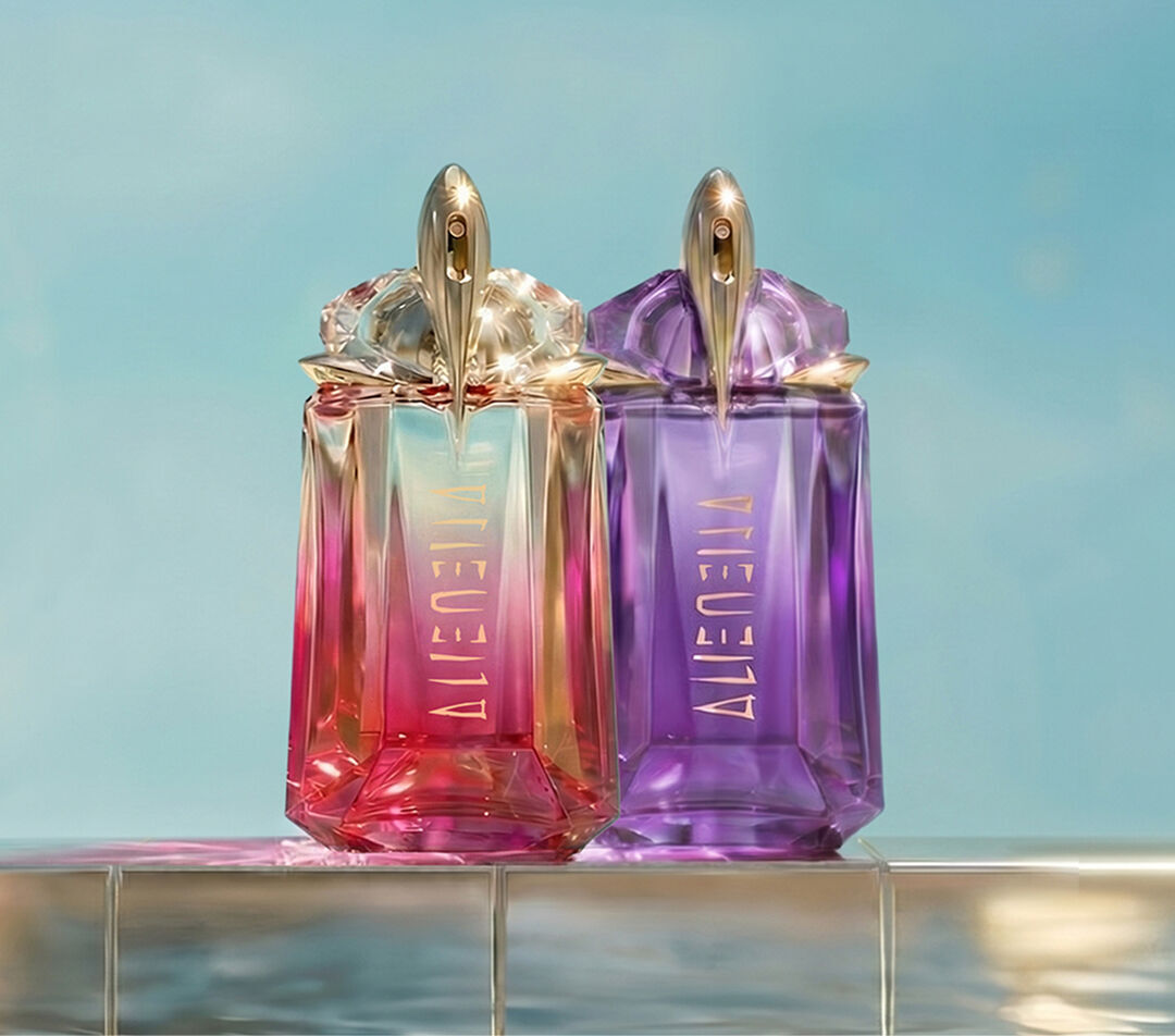 Two Mugler Alien Eau de Parfum and Alien Pulp perfume bottles in pink and violet stand side by side on a tiled pool edge with water reflections and a soft blue backdrop.
