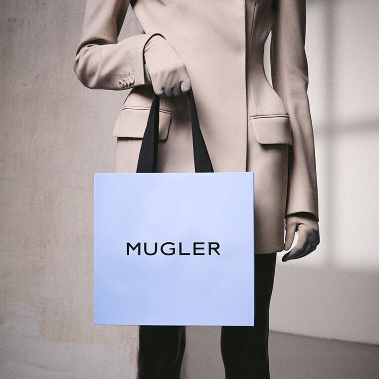 A person wearing a tailored Mugler blazer and gloves holding a light blue Mugler shopping bag with black handles, representing luxury gifting.