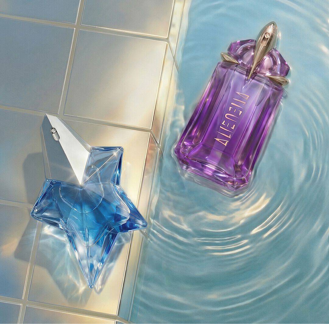 Mugler Angel Eau de Parfum shaped perfume bottle beside a purple Mugler Alien Eau de Parfum bottle floating in shimmering pool water next to pale tiles.