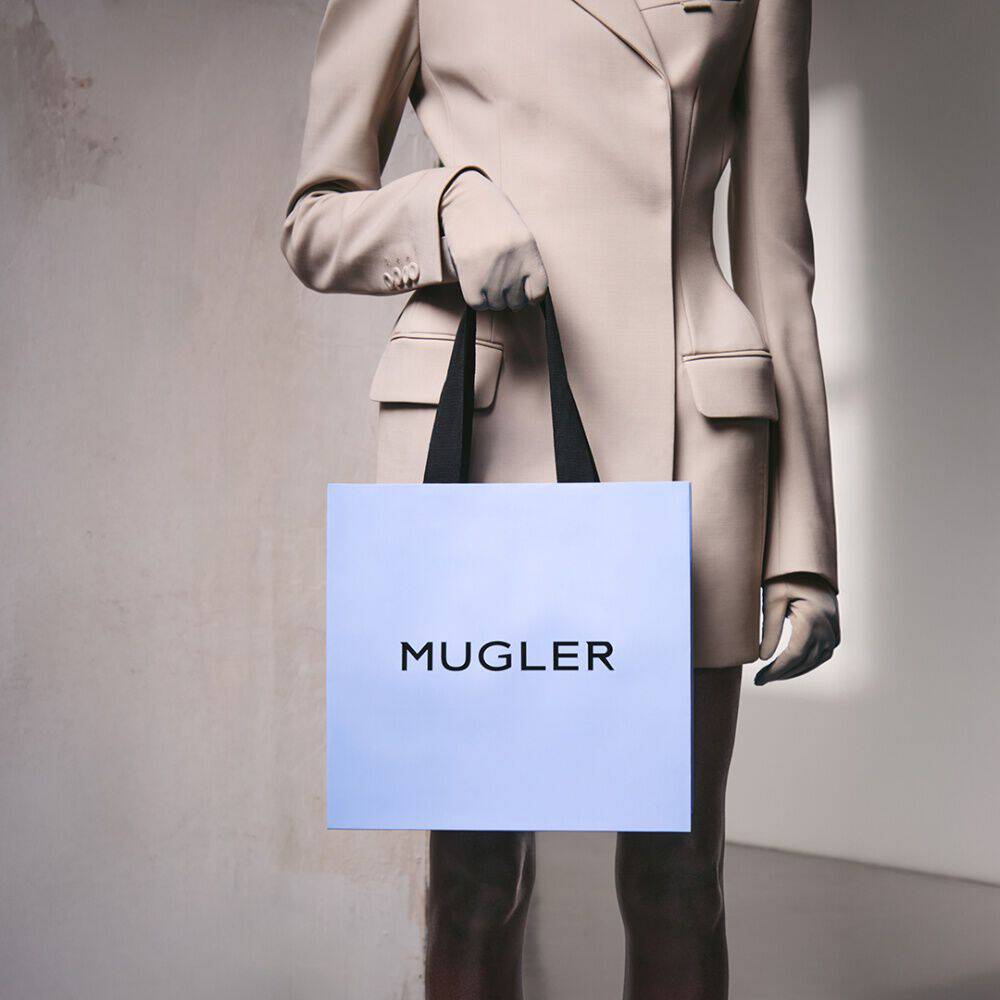 Person in beige blazer holding a light blue Mugler shopping bag with black handles against a neutral wall