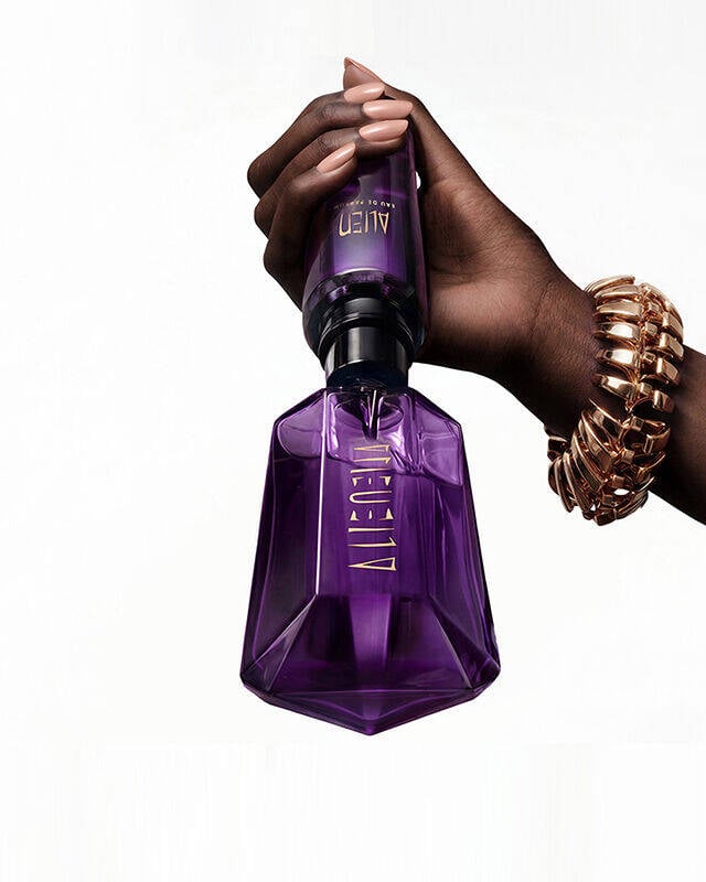 A hand wearing a gold chain-link bracelet refilling a purple Alien Eau de Parfum bottle with an official purple refill bottle at home, featuring the text overlay "refill at home".