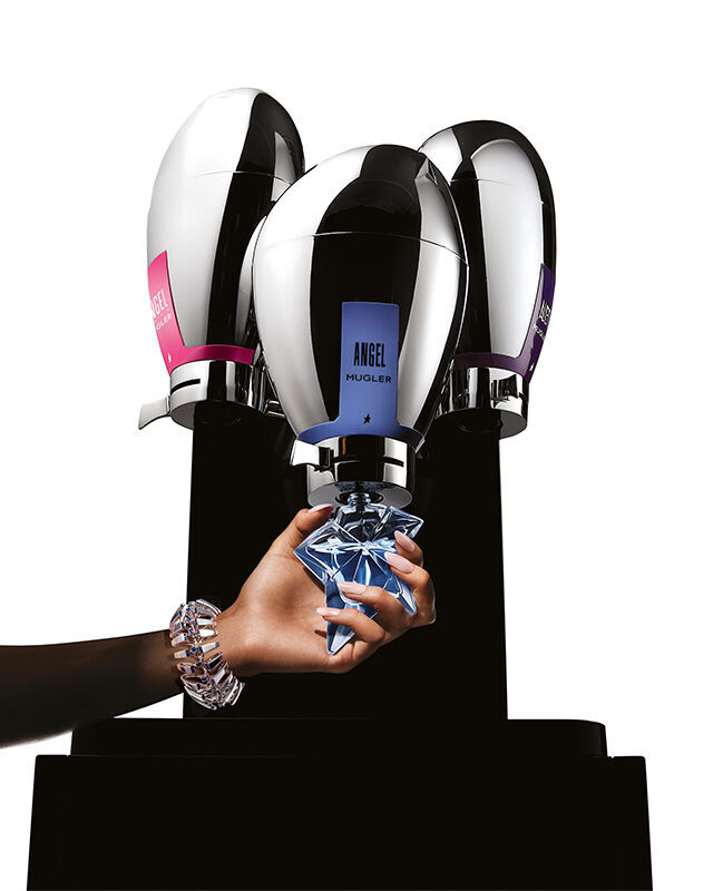 A sleek, futuristic metallic three-nozzle Mugler fragrance fountain dispenser with a hand refilling a glass bottle underneath.