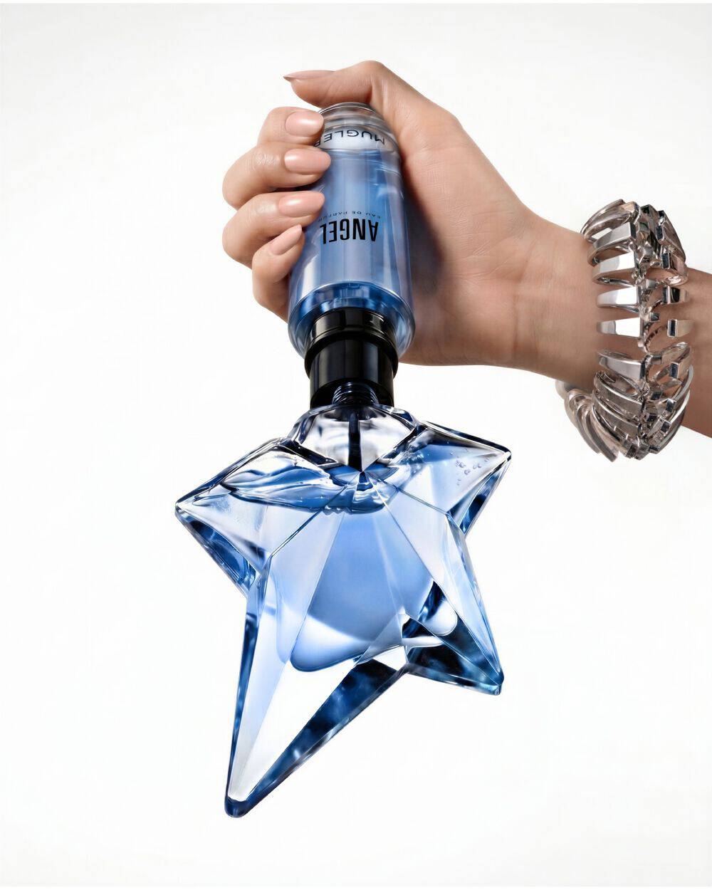 Close-up of a hand refilling the iconic blue star-shaped Angel Eau de Parfum bottle using a blue Mugler refill bottle, set against a soft beige background with a partial view of a model's face.