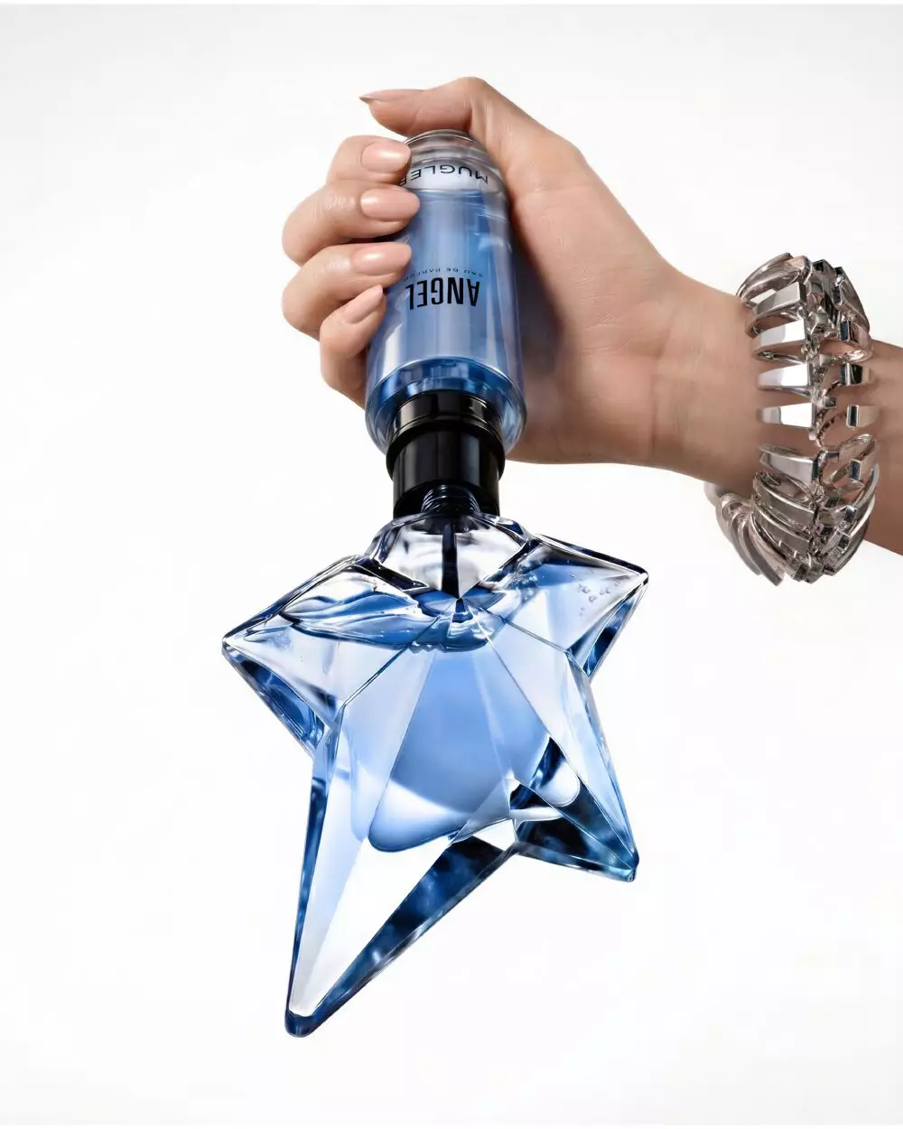 Close-up of a hand refilling the iconic blue star-shaped Angel Eau de Parfum bottle using a blue Mugler refill bottle, set against a soft beige background with a partial view of a model's face.
