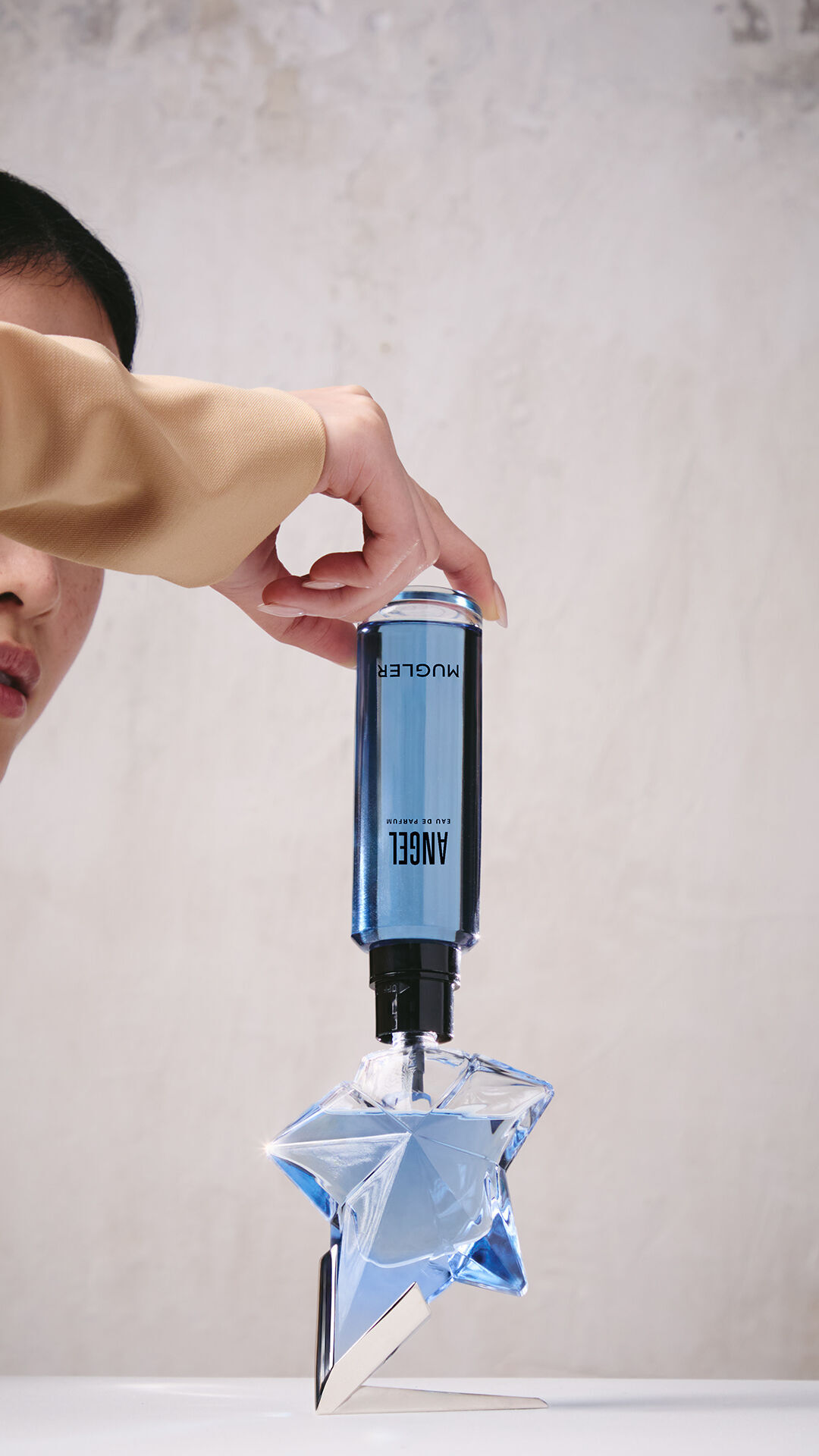 A hand refilling the blue star-shaped Angel Eau de Parfum bottle using a blue refill bottle upside down.