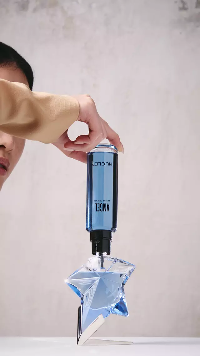 A hand refilling the blue star-shaped Angel Eau de Parfum bottle using a blue refill bottle upside down.