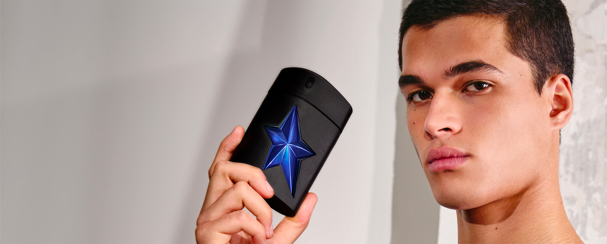 Young male model holding Mugler A*Men Stellar black cylindrical bottle with electric blue star, masculine luxury fragrance lifestyle shot